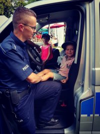 Obraz przedstawia policjanta Posterunku Policji w Ciechanowcu  na imprezie plenerowej w Ciechanowcu rozmawiającego  z dziećmi