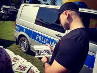 Obraz przedstawia mieszkańca miasta Ciechanowiec zapoznającego się z ulotką promocyjną dotyczącą zawodu policjanta na tle radiowozu