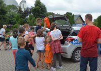 Obraz przedstawia policjantkę  z uczestnikami obozu w Hodyszewie, która pokazuje dzieciom i młodzieży  sprzęt policyjny oraz radiowóz