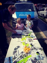 Na zdjęciu policjant dzielnicowy rozmawia z dziećmi o szeroko rozumianym bezpieczeństwie na imprezie plenerowej w Kuleszach Kościelnych rozdając elementy odblaskowe. W tle widoczny radiowóz.