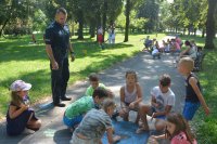 Obraz przedstawia dzieci uczestniczące na pikniku edukacyjnym wraz z policjantem, który prowadzi prelekcje dla dzieci