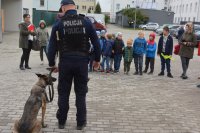 Obraz przedstawia policjanta pokazującego dzieciom psa służbowego