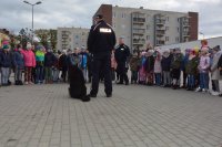 Obraz przedstawia policjanta przewodnika opowiadającego dzieciom na placu komendy o specyfice służby z psem