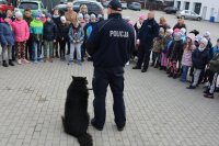 Obraz przedstawia policjanta przewodnika opowiadającego dzieciom na placu komendy o specyfice służby z psem