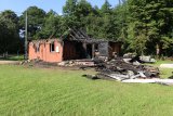 na pierwszym planie widać trawnik na drugim drewniany dom który uległ częściowemu spaleniu spalona więźba dachowa i ściany dookoła domu leżą spalone resztki ścian
