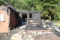 na pierwszym planie widać trawnik na drugim drewniany dom który uległ częściowemu spaleniu spalona więźba dachowa i ściany dookoła domu leżą spalone resztki ścian