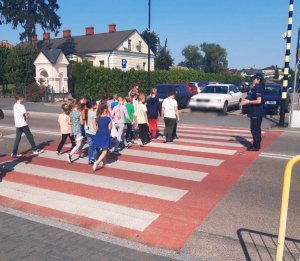 Obraz przedstawia policjantkę na spotkaniu z dziećmi przechodzącymi przez przejście dla pieszych
