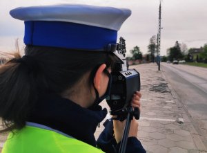 Obraz przedstawia policjantkę wydziału ruchu drogowego stojącą tyłem dokonującą pomiaru prędkości. W tle widać jadące pojazdy