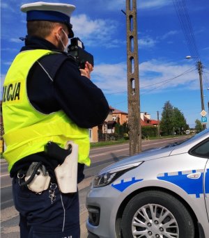 Obraz przedstawia policjanta Wydziału Ruchu Drogowego dokonującego pomiaru prędkości. W tle widać radiowóz