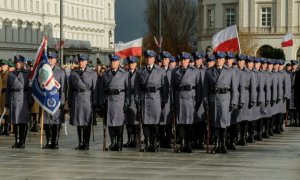 Obraz przedstawia policjantów stojących w szeregach. W tle widać flagi Polski