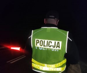 Obraz przedstawia policjanta w kamizelce odblaskowej zatrzymującego pojazd nocą