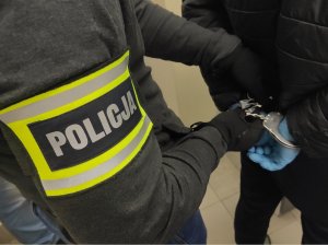 Obraz przedstawia ramie funkcjonariusza z opaską z napisem policja oraz dłonie zatrzymanego w rękawiczkach  i kajdankach