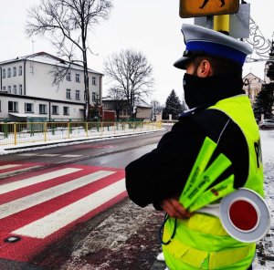 Obraz przedstawia policjanta stojącego przy przejściu dla pieszych trzymającego odblaski