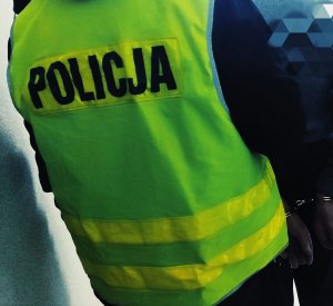 Obraz przedstawia policjanta stojącego tyłem  w kamizelce odblaskowej z zatrzymanym