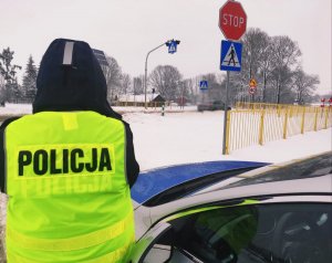 Obraz przedstawia policjanta stojącego przy radiowozie W tle widać przejeżdżające pojazdy oraz znaki drogowe