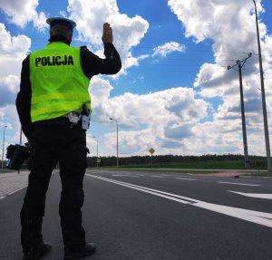obraz przedstawia policjanta trzymającego w jednej ręce urządzenie do pomiaru prędkości, a drugą trzyma w górze