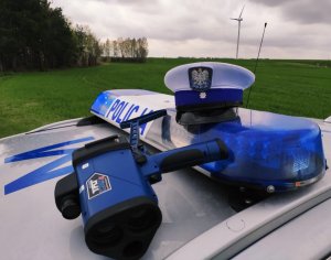Obraz przedstawia leżącą na radiowozie czapkę policjanta oraz urządzenie do pomiaru prędkości