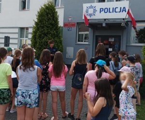 obraz przedstawia policjantkę z dziećmi na tle budynku Komendy Powiatowej Policji w Wysokiem Mazowieckiem