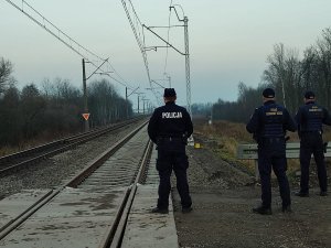 obraz przedstawia policjanta i strażników ochrony kolei na tle przejazdu kolejowego