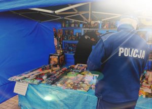 obraz przedstawia policjanta na tle stoiska ze sprzedażą materiałów pirotechnicznych