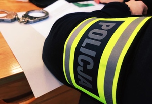 obraz przedstawia ramię policjanta z opaską z napisem policja, w tle kajdanki