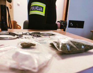 obraz przedstawia zabezpieczone narkotyki, w tle policjant z opaską na ramieniu z napisem policja
