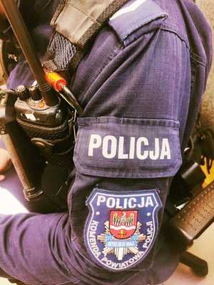 obraz przedstawia ramię policjanta z naszywką z napisem policja, w tle urządzenia do utrzymywania łączności