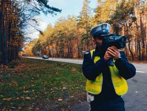 obraz przedstawia policjanta trzymającego urządzenie do pomiaru prędkości na tle masywu leśnego