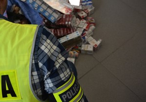obraz przedstawia ramię policjanta z opaską z napisem policja, w tle zabezpieczone papierosy
